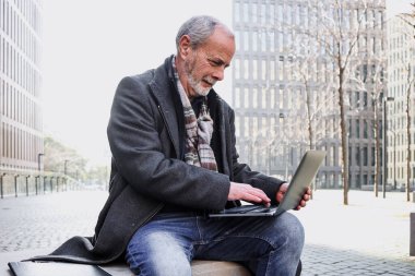 Hispanik kıdemli işadamı dizüstü bilgisayarını kullanarak kentin dışında bankta oturuyor, olgun bir adam bilgisayarla çalışıyor.