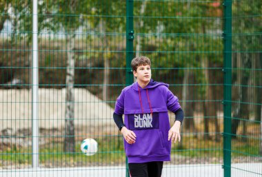 Menekşe kapüşonlu şirin bir genç basketbol oynuyor. Top öğrenme yeteneği olan genç bir çocuk şehir sahasında top sürüyor ve ateş ediyor. Çocuklar için hobi, aktif yaşam tarzı.