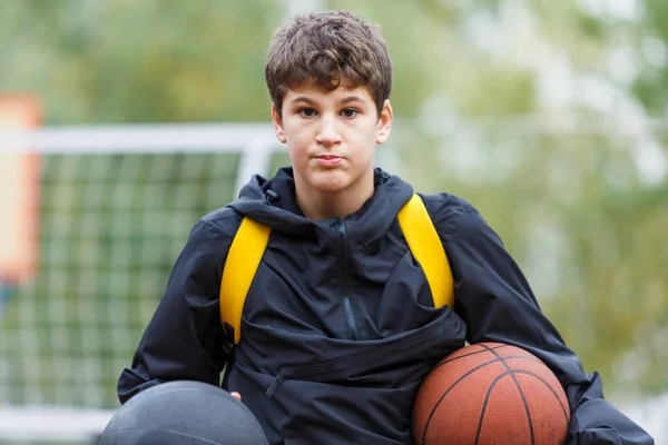 Cute young boy plays basketball on street playground. Teenager with orange basketball ball outside. Hobby, active lifestyle, sport activity for kids.