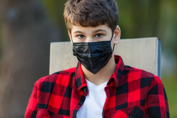 顔に黒い保護マスクをしたかわいい少年を閉じます コロナウイルス 感染から保護するために医療マスクを身に着けている赤いチェックフランネルシャツの十代の若者 街中の隔離 — ストック写真