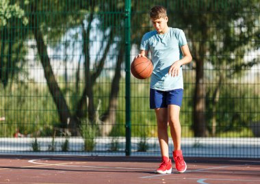 Tatlı genç bir çocuk yaz mevsiminde sokak bahçesinde basketbol oynar. Dışarıda turuncu basketbol topu olan yeşil tişörtlü bir genç. Hobi, aktif yaşam tarzı, çocuklar için spor aktiviteleri.