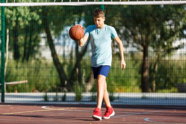 Tatlı genç bir çocuk yaz mevsiminde sokak bahçesinde basketbol oynar. Dışarıda turuncu basketbol topu olan yeşil tişörtlü bir genç. Hobi, aktif yaşam tarzı, çocuklar için spor aktiviteleri.