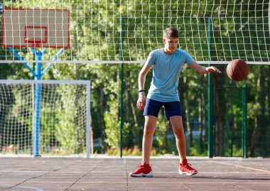 Tatlı genç bir çocuk yaz mevsiminde sokak bahçesinde basketbol oynar. Dışarıda turuncu basketbol topu olan yeşil tişörtlü bir genç. Hobi, aktif yaşam tarzı, çocuklar için spor aktiviteleri.