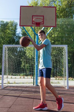 Tatlı genç bir çocuk yaz mevsiminde sokak bahçesinde basketbol oynar. Dışarıda turuncu basketbol topu olan yeşil tişörtlü bir genç. Hobi, aktif yaşam tarzı, çocuklar için spor aktiviteleri.