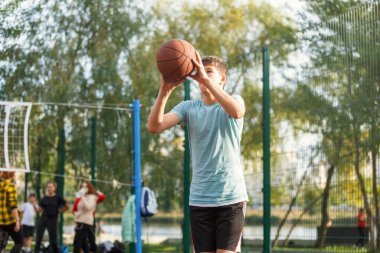 Tatlı genç bir çocuk yaz mevsiminde sokak bahçesinde basketbol oynar. Dışarıda turuncu basketbol topu olan yeşil tişörtlü bir genç. Hobi, aktif yaşam tarzı, çocuklar için spor aktiviteleri.