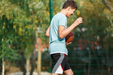 Tatlı genç bir çocuk yaz mevsiminde sokak bahçesinde basketbol oynar. Dışarıda turuncu basketbol topu olan yeşil tişörtlü bir genç. Hobi, aktif yaşam tarzı, çocuklar için spor aktiviteleri.