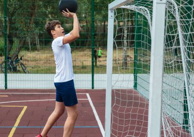 Tatlı genç bir çocuk yaz mevsiminde sokak bahçesinde basketbol oynar. Dışarıda turuncu basketbol topu olan beyaz tişörtlü bir genç. Hobi, aktif yaşam tarzı, çocuklar için spor aktiviteleri.