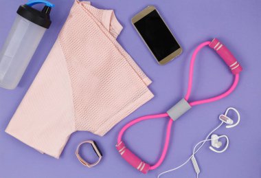 Menekşe arka plandaki kadınlar için spor kıyafetleri ve aksesuarlar. Tişörtlü spor modası, elastik bantlar, kulaklıklar, telefon, şişe. Sağlıklı, aktif yaşam tarzı konsepti