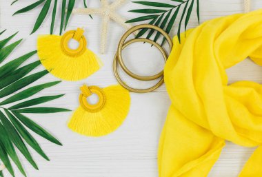 Tropik palmiye yaprakları, hasır şapka, denizyıldızı, beyaz ahşap arka planda deniz kabukları. Modayı takip eden sarı kıyafetler ve aksesuarlar. Düz yat, kapat. Yaz, tatil, tatil konsepti.  