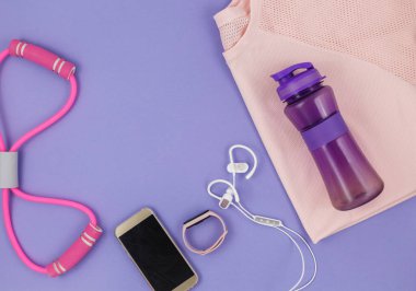 Menekşe arka plandaki kadınlar için spor kıyafetleri ve aksesuarlar. Tişörtlü spor modası, elastik bantlar, kulaklıklar, telefon, şişe. Sağlıklı, aktif yaşam tarzı konsepti