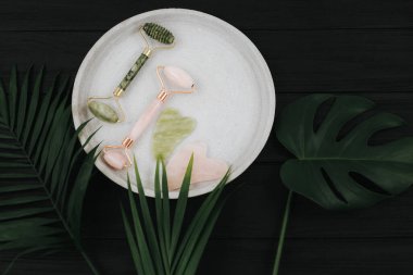 Kristal gül kuvars ve masaj aleti yeşim Gua sha yüz silindiri siyah arka planda, palmiye yaprakları ile tepside. Anti-yaş masajı için aletler, evde doğal yüz gerdirme. Karanlık ruh hali tarzı