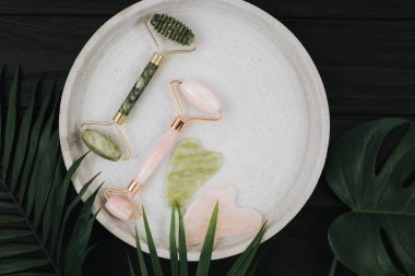 Kristal gül kuvars ve masaj aleti yeşim Gua sha yüz silindiri siyah arka planda, palmiye yaprakları ile tepside. Anti-yaş masajı için aletler, evde doğal yüz gerdirme. Karanlık ruh hali tarzı