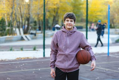 Turuncu basketbol topu olan sevimli ergen bahar-yaz döneminde sokak parkında basketbol oynuyor. Hobi, aktif yaşam tarzı, çocuklar için spor aktiviteleri.