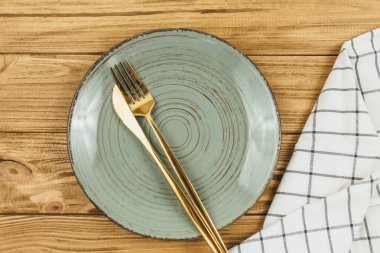 Çatallı yeşil boş tabak, tahta masanın üzerinde beyaz çizgili bıçak peçetesi. Altın çatal bıçak takımı, sofra takımı.