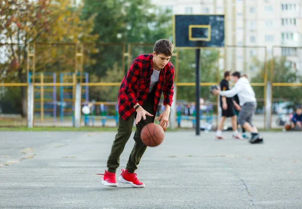 A cute young boy plays basketball on a street playground. Teenager in red flannel checked shirt with orange basketball ball outside. Hobby, active lifestyle, sports activity for kids.