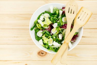 Keçi peynirli taze yeşil salata, ahşap masadaki büyük beyaz kasede yumurtalar. Yeşil yapraklı vejetaryen salatası ve tahta kaşıklı sebzeler. Üst Manzara. Sağlıklı vejetaryen yemekleri. Kapat.