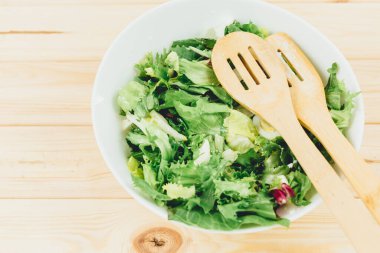 Keçi peynirli taze yeşil salata, ahşap masadaki büyük beyaz kasede yumurtalar. Yeşil yapraklı vejetaryen salatası ve tahta kaşıklı sebzeler. Üst Manzara. Sağlıklı vejetaryen yemekleri. Kapat.