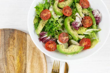 Avokadolu taze yeşil salata, domates, tahta tepside büyük beyaz bir kasede keçi peyniri. Yeşil yapraklı ve sebzeli vejetaryen salatası. Gri masadaki üst manzara. Sağlıklı vejetaryen yemekleri. Kapat.