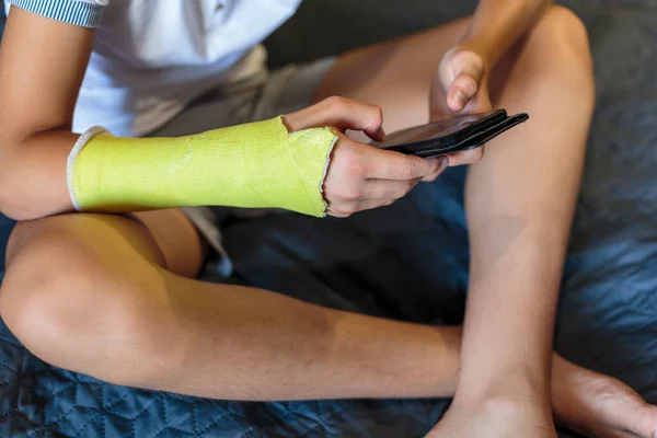 Boy with broken hand relaxing. Close up young handsome Teenage with yellow plaster playing smartphone at home.