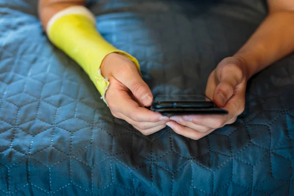 Boy with broken hand relaxing. Close up young handsome Teenage with yellow plaster playing smartphone at home.