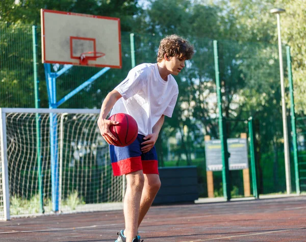 Cute teenager plays basketball at a city playground. A boy holds a basketball ball in his hands outside. Active life, hobby, sports for children