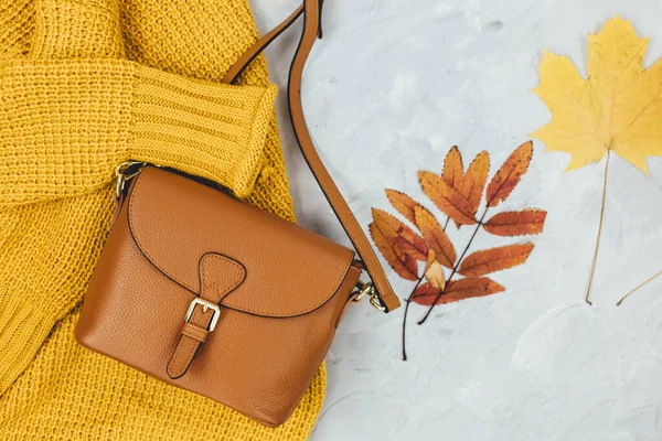 Orange leather women bag, knitted sweater, autumn leaves on gray background. Trendy autumn accessories. Cozy outfit