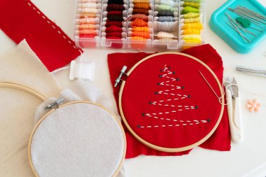 Kırmızı bir kumaş, şenlikli bir Noel ağacı tasarımının yer aldığı ahşap işlemeli bir halka şeklinde gerilir. İğneler ve iplikler etrafa saçılmış, rahat bir zanaat atmosferini vurguluyorlar..