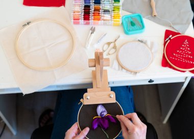 Kadın, el sanatları masasında ipek kurdele kullanarak mor bir çiçeği ustalıkla dikiyor. Parlak renkler ve araçlar nakış sanatına ve yaratıcılığa bir adanmışlık göstermektedir..