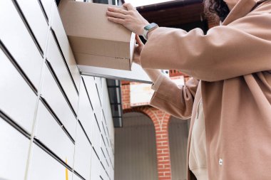 Bir kadın güvenli bir teslimat kutusundan bir paket çıkarıyor. O, bileğine bir akıllı seğirme takar. Bu sahne güneşli bir günde, bir yerleşim bölgesinde geçiyor..