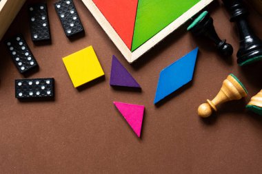 Kahverengi ahşap bir yüzey üzerinde çeşitli masa oyunları ve oyun parçaları düzenlenir. Renkli yapboz parçaları, domino taşları, dama ve satranç seti içeriyor..