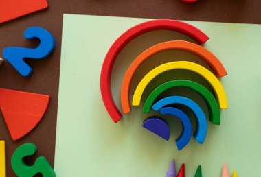 Parlak renkli ahşap gökkuşağı parçaları yeşil bir kağıt üzerinde sanatsal olarak düzenlenir. Çevrelerinde çeşitli renkli şekiller ve sayılar var. Canlı bir oyun sahnesi yaratıyorlar..