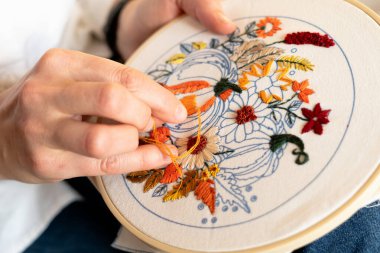 Bir kişi, balkabakları ve canlı çiçeklerin yer aldığı nakışlı bir proje üzerinde çalışıyor. Bu yaratıcı etkinlik, kapalı bir alanda, yumuşak bir kumaş arka planda, sanatsal beceriler sergiliyor..