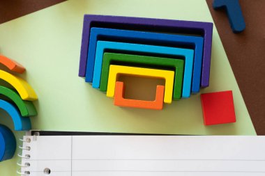 Parlak renkli ahşap şekiller kahverengi bir masanın üstüne düzgünce istiflenmiştir. Yakınlarda, boş bir defter evde ya da okulda çocuklar için yaratıcı oyun ve eğitim aktivitelerini davet eder.