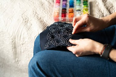 Kadın bir sandalyede oturmuş iğne iplik kullanarak koyu renk kumaş üzerine dikiş dikiyor. Yanlarındaki konteynırda renkli iplikler depolanmış. İçerideki ortam çok rahat..