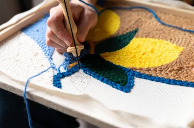 Genç bir kadın, el yapımı bir tuvalde kanca kullanarak renkli bir tekstil sanat eseri yaratıyor. Zanaat çeşitli iplik renkleriyle detaylı bir çalışma içeriyor. Mekan iyi aydınlatılmış..