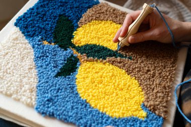 Bir el, tuval üzerinde renkli halı tasarımları yapmak için bir kanca aleti tutar. Konferansta ayrıntılı çalışma ve kapalı alandaki parlak iplik üzerinde duruluyor. El, kapalı alanda el yapımı oturum sırasında halı tasarımı oluşturmak için kanca kullanır