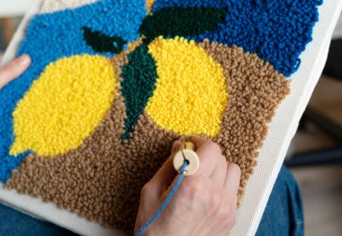 Kadın, mavi, yeşil ve sarı renkli bir halı yapmak için kanca kullanıyor. Dekorun yumuşak bir dokusu var ve kapalı alanda yapılırken ayrıntılı desenleri var..