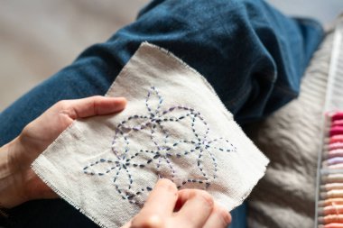 Sandalyeye oturmuş iğne iplik kullanarak beyaz kumaşa dikiş dikiyor. Yanlarındaki konteynırda renkli iplikler depolanmış. İçerideki ortam çok rahat..