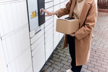 Bulutlu bir günde bir binanın önünde dikilirken elinde iki karton kutu ve bir telefon tutan kadın.