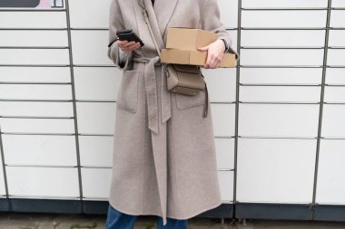 Gün boyunca dolapları olan bir duvarın önünde cep telefonu ve kutu tutan bir kadın.