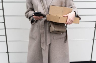 Kadın dışarıda dolapların önünde duruyor. Bir elinde cep telefonu, diğer elinde kutu tutuyor. Bir ceket giyiyor ve yanında küçük bir çanta var..