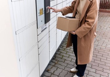 Bir elinde iki karton kutu, diğer elinde bir cep telefonu olan biri dışarıda duruyor. Açık renk bir ceket giyiyorlar ve binanın girişinin yakınındalar..