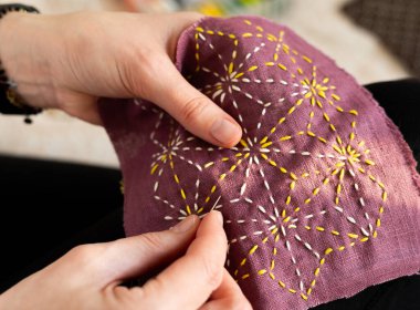 Bir insan mor bir kumaş parçasındaki sarı ve beyaz iplikleri kullanarak bir tasarımı dikmeye odaklanır. Dekor, doğal ışıkla dolu kapalı bir alan. Dikiş onarılıyor