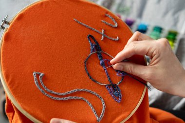 Kadın elinde iğne ve iplik tutarken turuncu bir kumaşa tasarım dikiyor. İplik makaraları arka planda görünür. Bu aktivite iyi ışık alan yaratıcı bir alanda gerçekleşiyor. İnsan turuncu kumaşa renkli iplikler diker