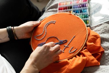 Kadın elinde iğne ve iplik tutarken turuncu bir kumaşa tasarım dikiyor. İplik makaraları arka planda görünür. Bu etkinlik, iyi ışık alan yaratıcı bir alanda gerçekleşiyor..