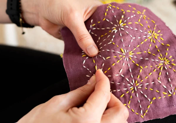 Kadının elleri mor bir kumaş parçasında sarı ve beyaz iplikler kullanarak bir tasarımı dikmeye odaklanıyor. Dekor, doğal ışıkla dolu kapalı bir alan..