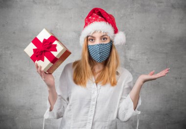 Güzel sarışın kadın Noel Baba şapkası takıyor. Şapka takıyor ve elinde kırmızı hediye kutusu var. Üzerinde kurdele var. Bir Corona - Covid 19 Noel konsepti. Stüdyo çekimi