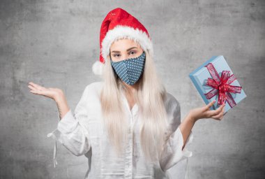 Noel hediyesi olan Noel Baba kadın gri arka planda izole edilmiş. Kırmızı Noel Baba şapkası takan ve hediye kutusunu tutan düşünceli bir genç kız. Bir Corona - Covid 19 Noel konsepti. Stüdyo çekimi