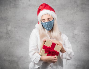 Gri arka planda izole edilmiş Noel Baba hediyeli bir kadın. Kırmızı Noel Baba şapkası takan ve hediye kutusunu tutan mutlu bir genç kız. Bir Corona - Covid 19 Noel konsepti. Stüdyo çekimi