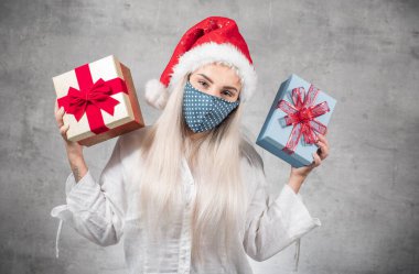 Bir Corona - Covid 19 Noel konsepti. Noel Baba kadın gri arka planda izole edilmiş Noel hediyeleri tutuyor. Kırmızı Noel Baba şapkası takan ve hediye kutusunu tutan mutlu genç kız. Stüdyo çekimi